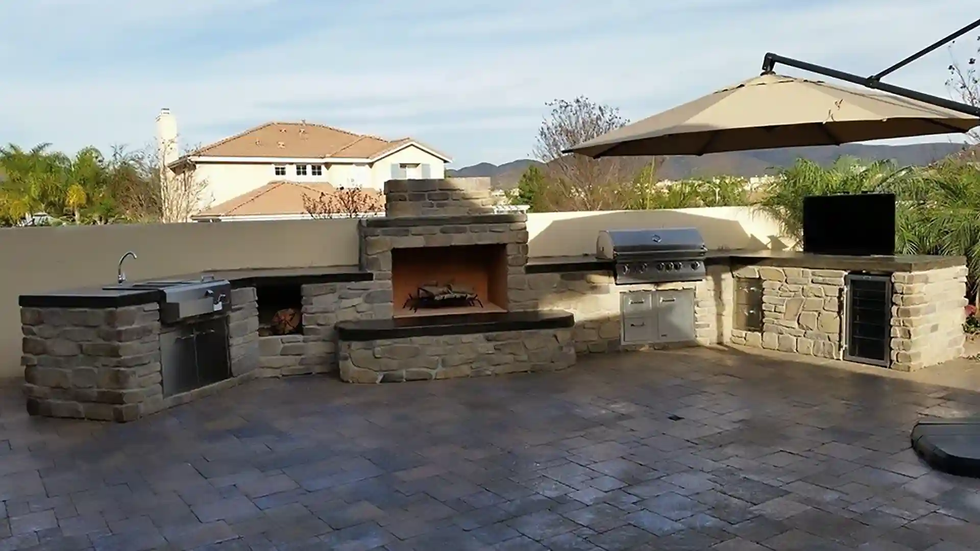 photo of an extended outdoor kitchen on a paver patio with a sink, cabinetry, grill, beverage fridge, outdoor fireplace, and more